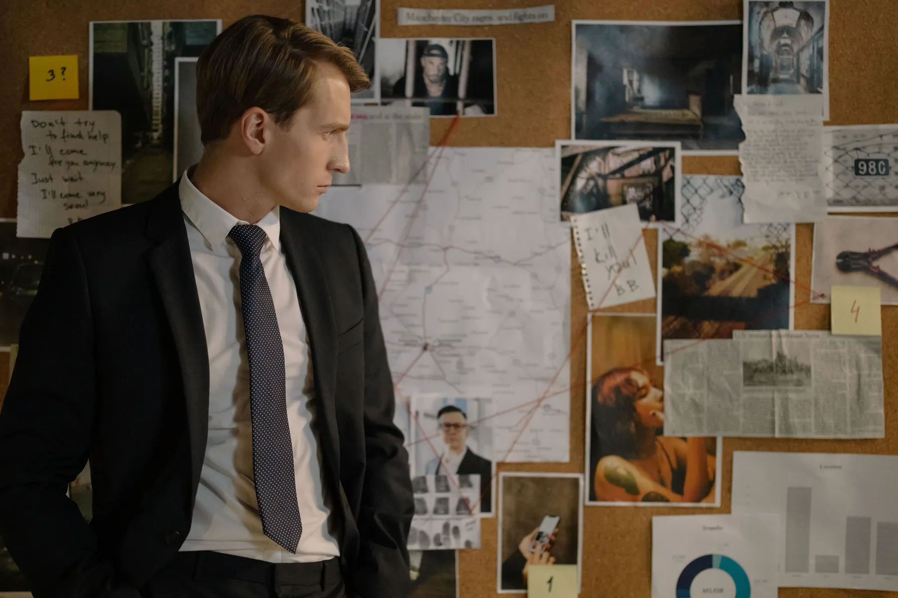 A man in a suit studies photographs, notes, maps, and red threads on an investigation board.